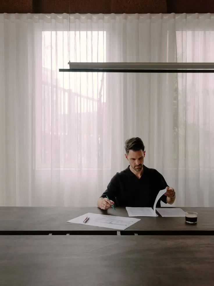 Een ondernemer leest papieren aan een lange tafel voor lichte gordijnen