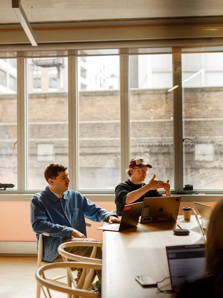 Twee collega's overleggen aan een tafel met laptops bij een groot raam